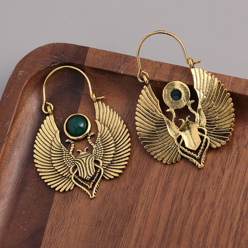 Gold-plated Egyptian goddess dangle earrings with winged design and green stone displayed on wooden tray