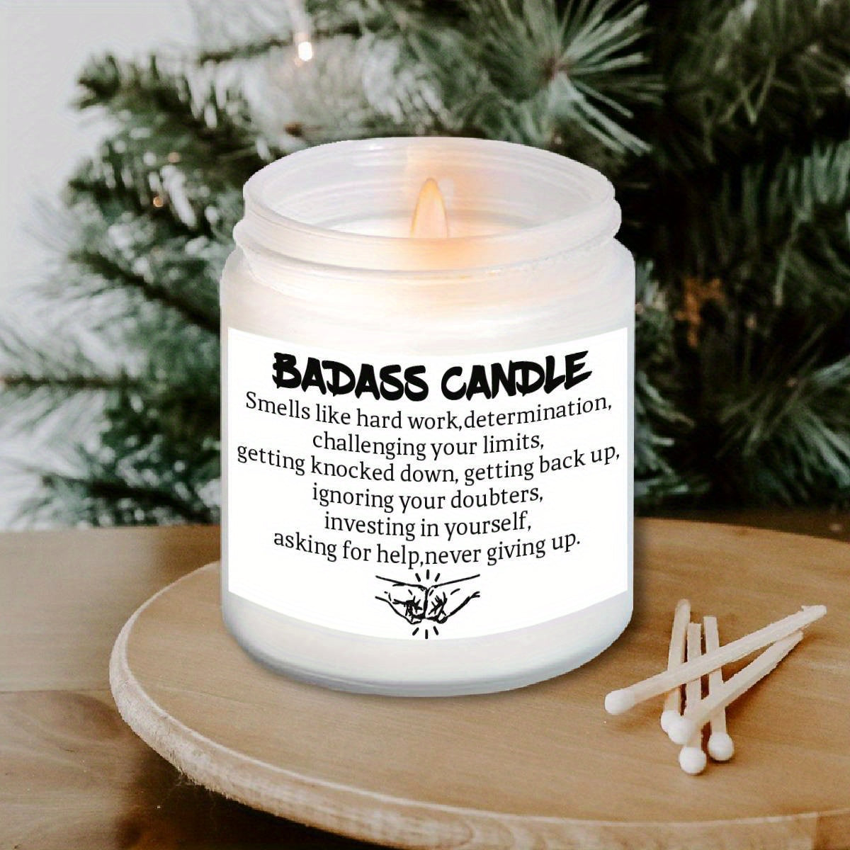 Humorous novelty candle in frosted glass jar with bold motivational quote label, lit on a wooden surface with greenery background