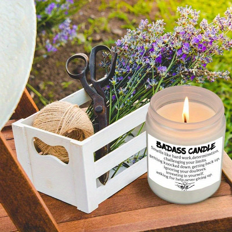 Humorous novelty candle in glass jar with bold quote label, lit on a wooden table beside lavender flowers and rustic decor accessories