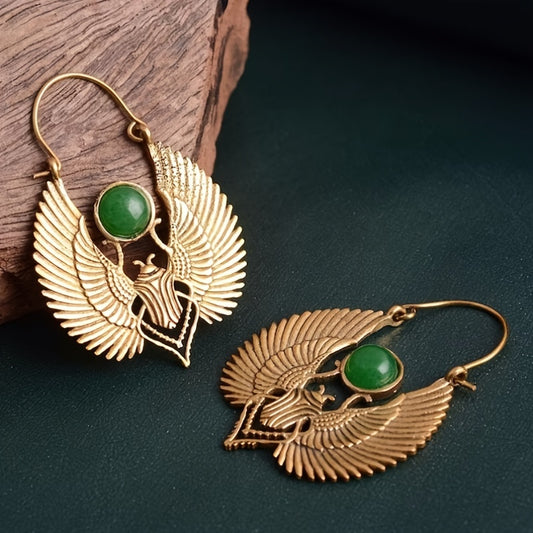 Gold-plated winged Egyptian goddess earrings with green stone accent on dark surface.