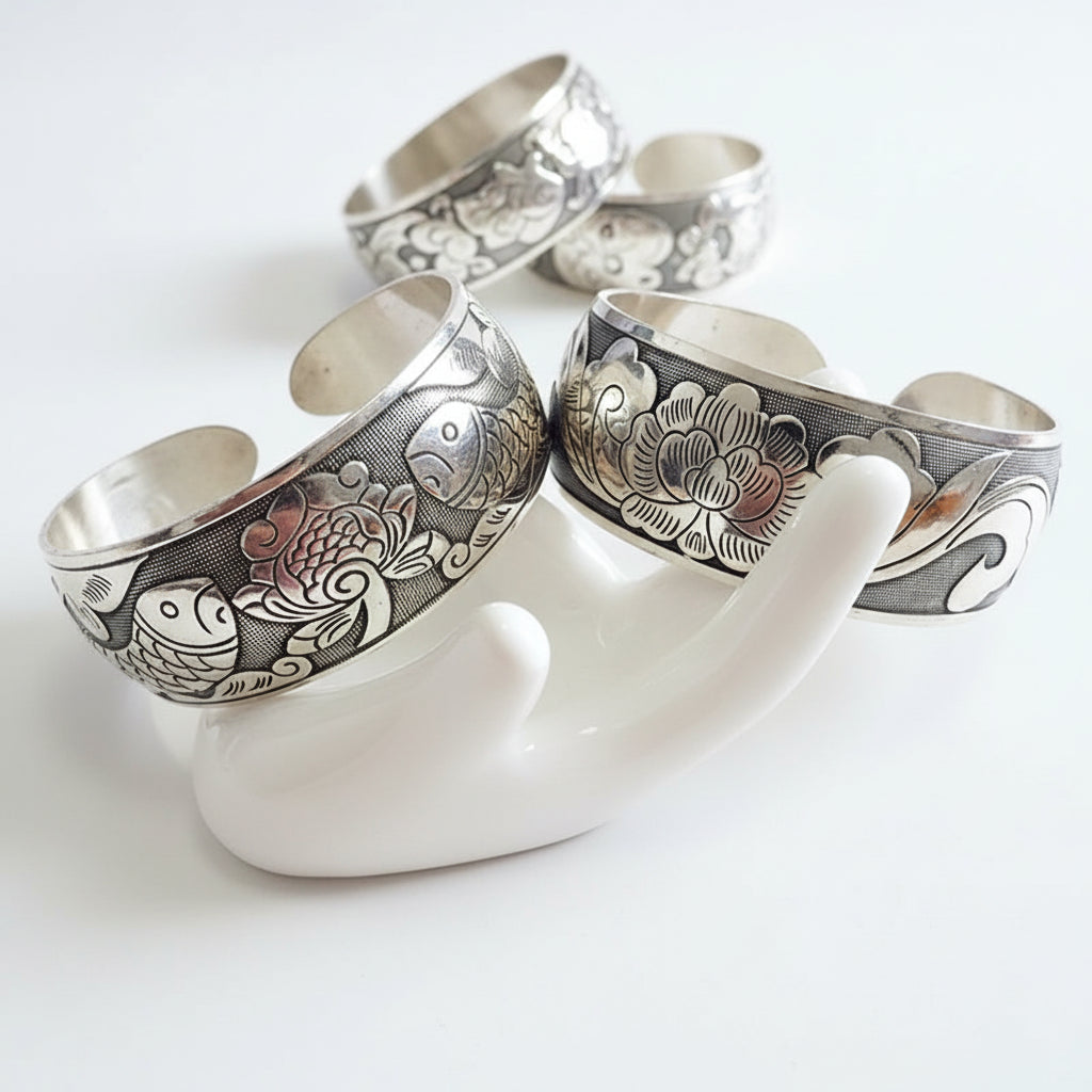 Two silver bangles with intricate designs on a white hand-shaped stand.