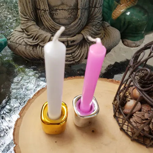 Two candles, one white and one pink, in decorative holders on a wooden surface with a statue and basket in the background. - Clover's Charm