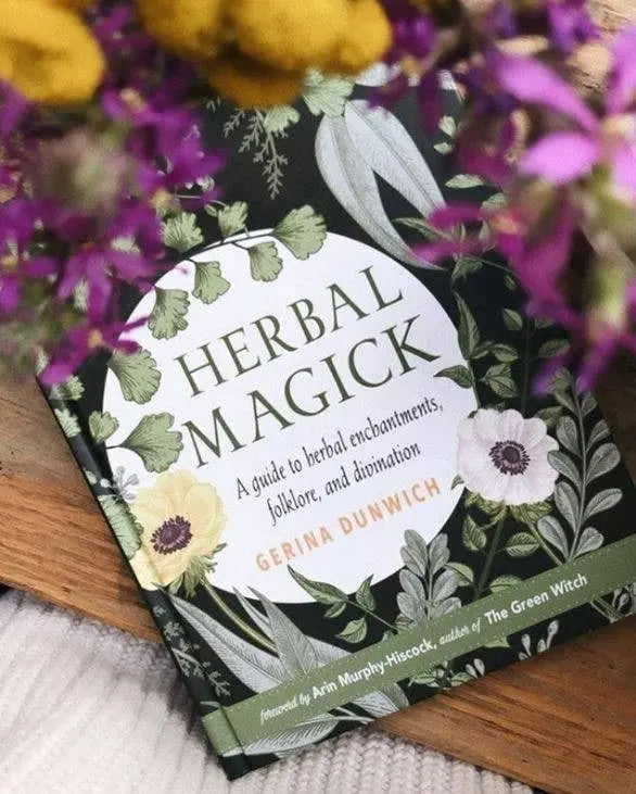Book titled 'Herbal Magick' by Gerina Dunwich surrounded by flowers on a wooden surface.