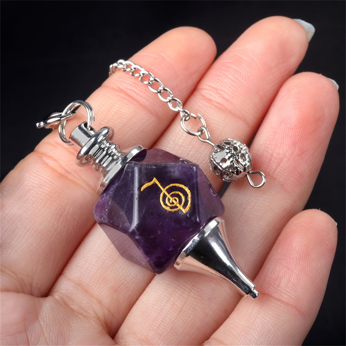 Purple pentagonal pendant with a black spike and silver chain held in a hand against a dark background