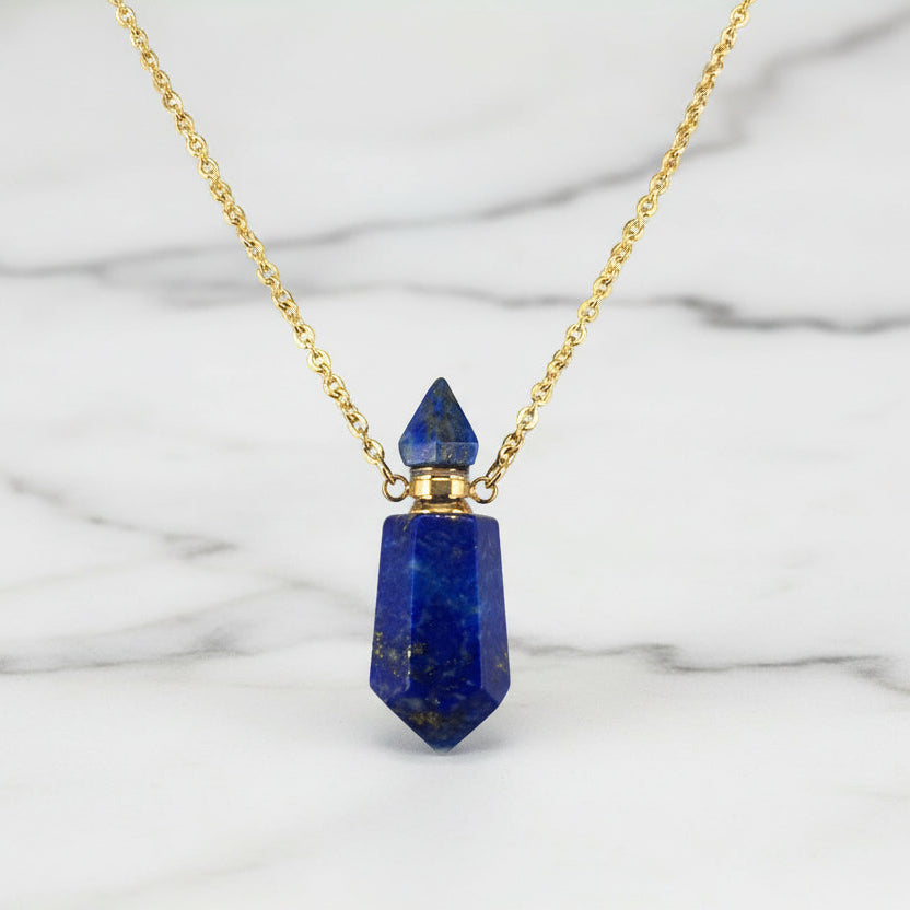 Natural blue gemstone prism perfume bottle necklace with gold plated stainless steel chain on a white marble surface.
