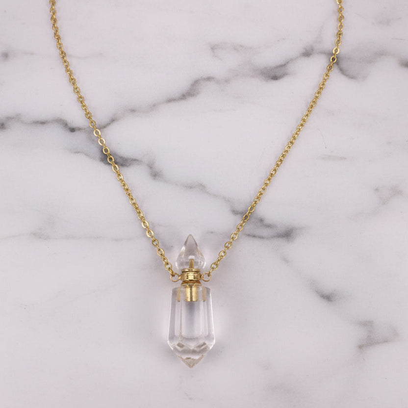 Natural clear gemstone prism perfume bottle necklace with gold plated stainless steel chain on a white marble surface.
