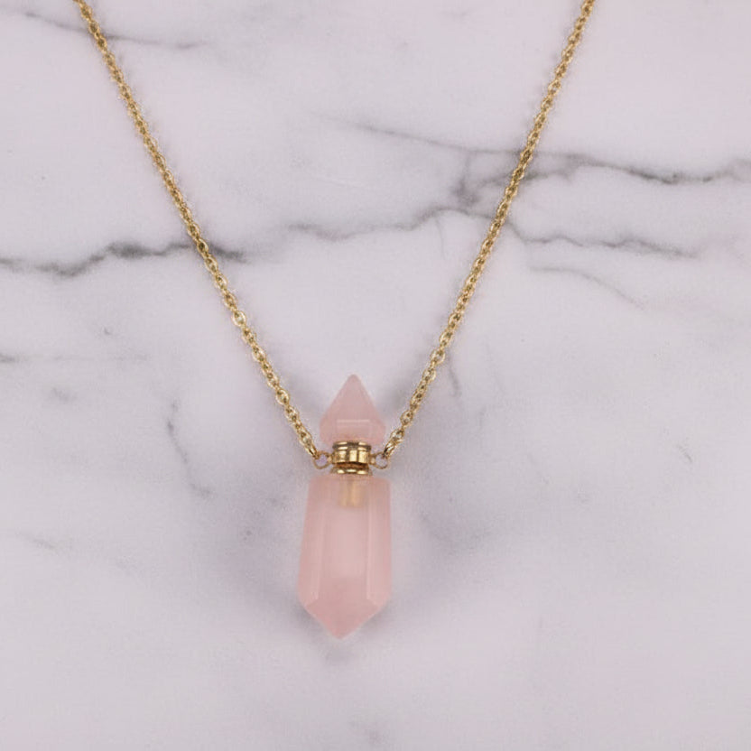 Natural pink gemstone prism perfume bottle necklace with gold plated stainless steel chain on a white marble surface.
