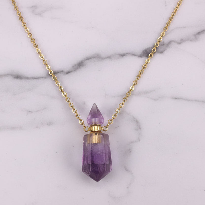 Natural purple gemstone prism perfume bottle necklace with gold plated stainless steel chain on a white marble surface.
