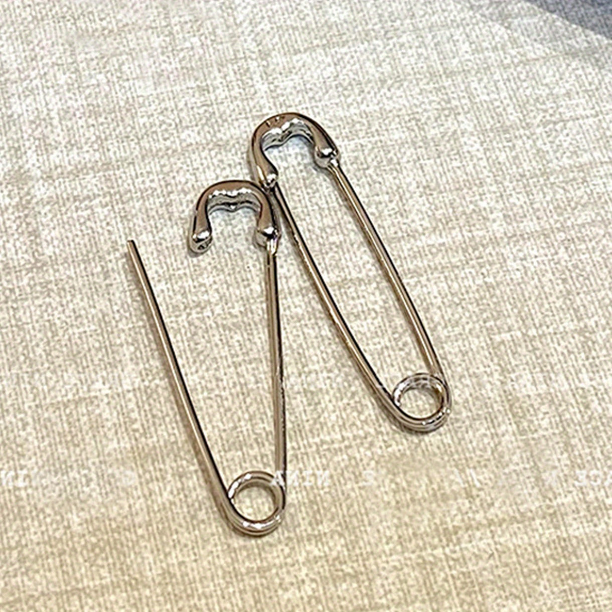 Two metal safety pins styled earrings on a textured beige fabric background